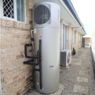 Heat pump water heater systen is installed outside a brick house, with black pipes connected to it.